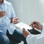 TRD Research represented by a person in a white coat holding a clipboard and talking to a patient.